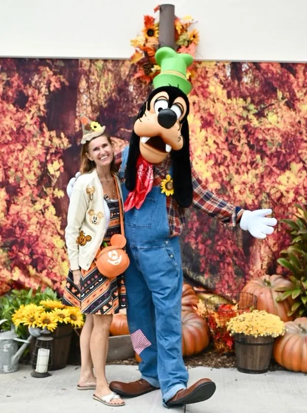 A guest posing with Goofy in front of the fall leaves and pumpkin display