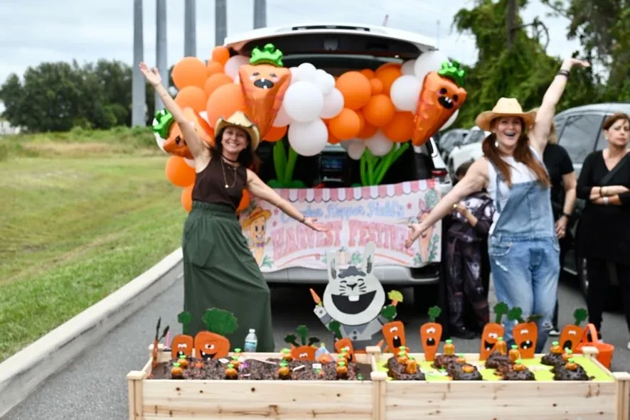 Two parents in overalls and straw hats with arms wide open at a carrot-themed Trunk or Treat garden bed