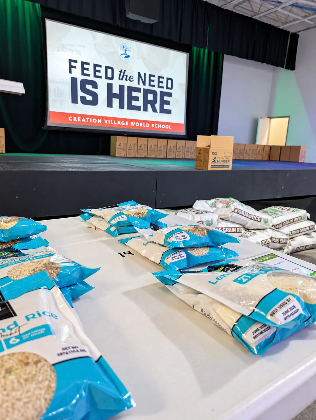 Sacks of rice and lentils ready for the Feed the Need packing day, with the Feed the Need Is Here screen